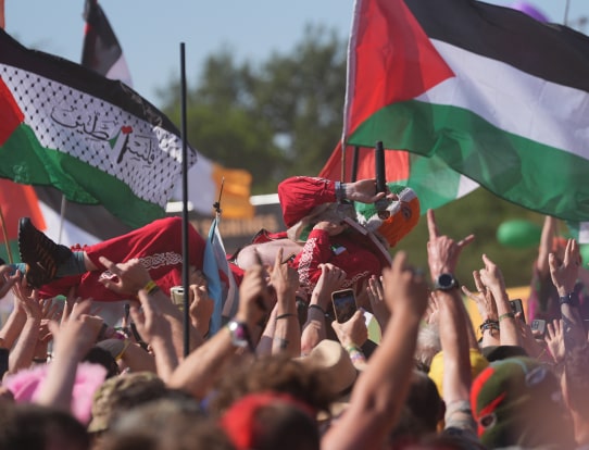 A crowd at a festival lifts a performer above their heads while waving flags, including Palestinian flags.