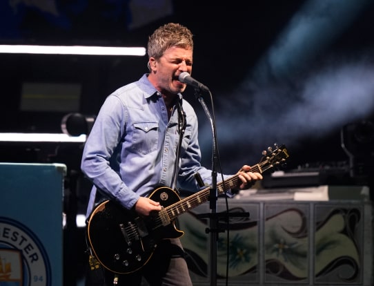 Noel Gallagher performing on stage, singing into a microphone while playing electric guitar.