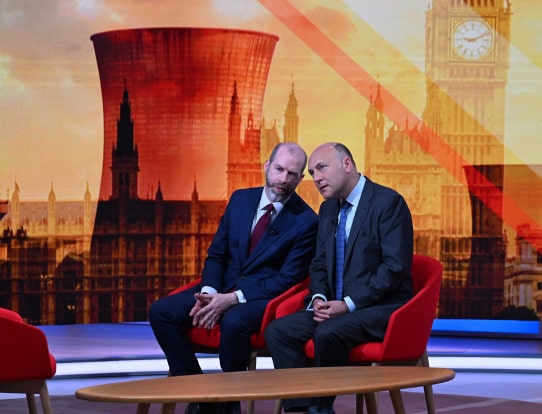Two men in suits sit on studio chairs, leaning in to talk during a televised discussion.
