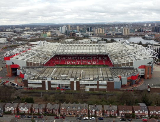Aerial view of Manchester United’s Old Trafford stadium with surrounding buildings and cityscape in the background.