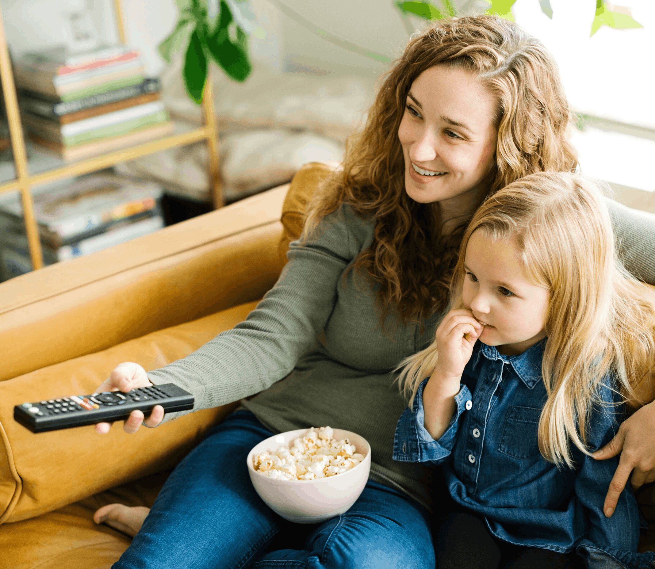 A woman and child on a cozy couch share a bowl of popcorn, watching TV with smiles. The room is warmly decorated with plants and books nearby.