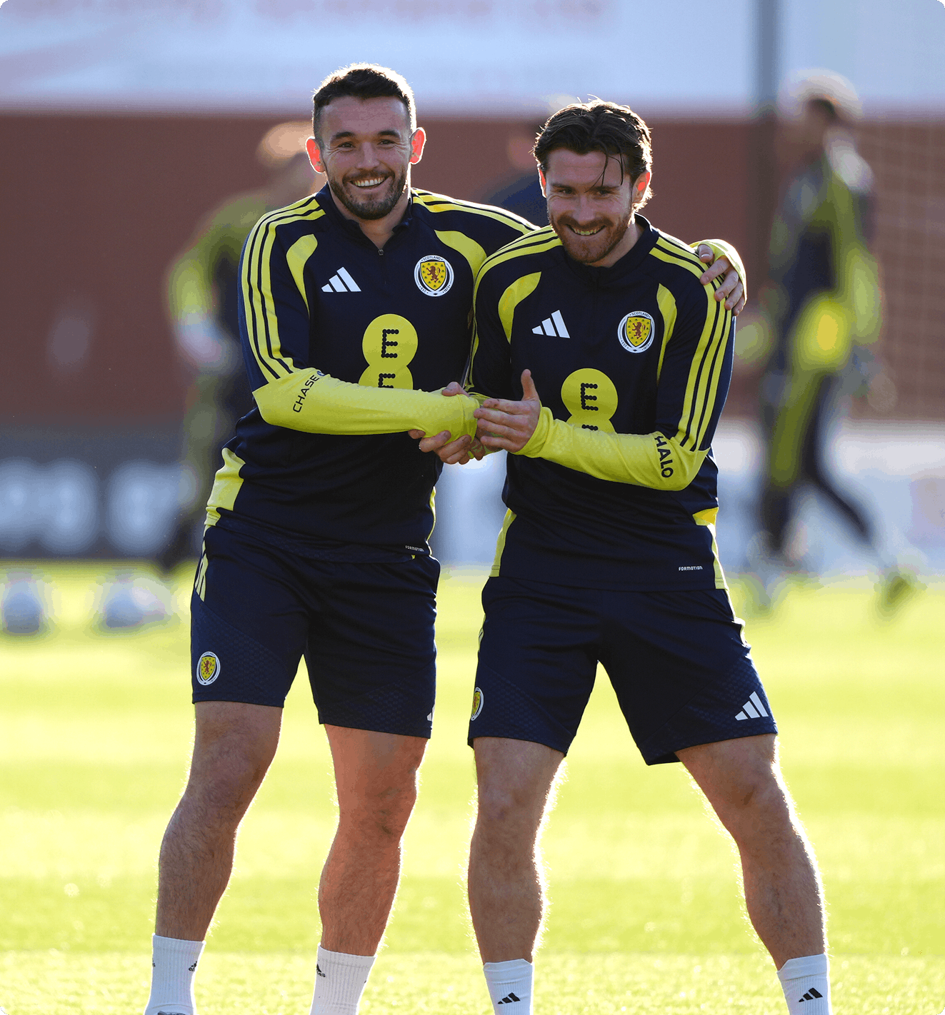 Two soccer players in navy and yellow Scotland training kits smile and clasp hands warmly on a sunlit field, conveying camaraderie and joy.