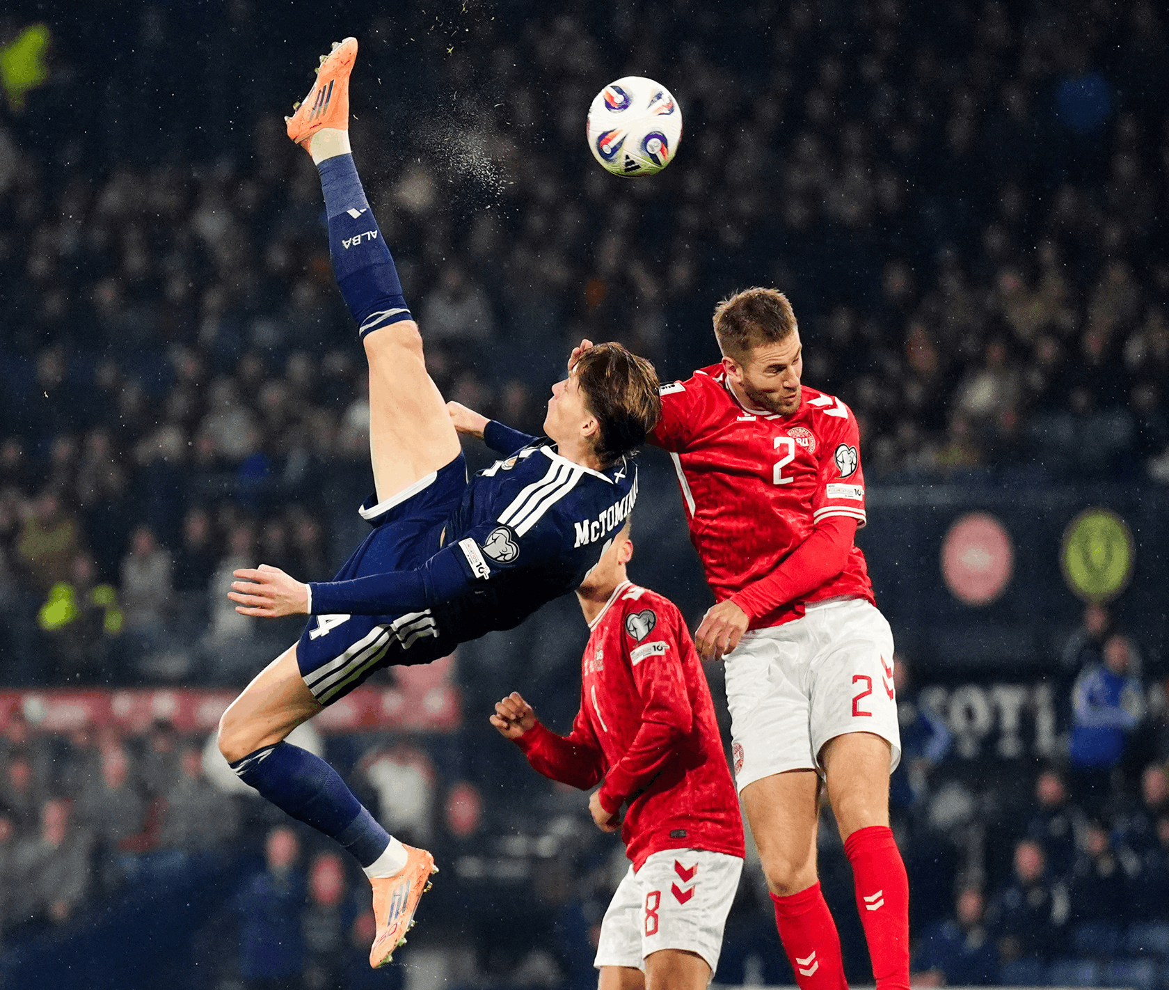 A soccer player in a blue jersey attempts a bicycle kick, while two players in red jerseys look on. The action is intense, with a crowd blurred in the background.