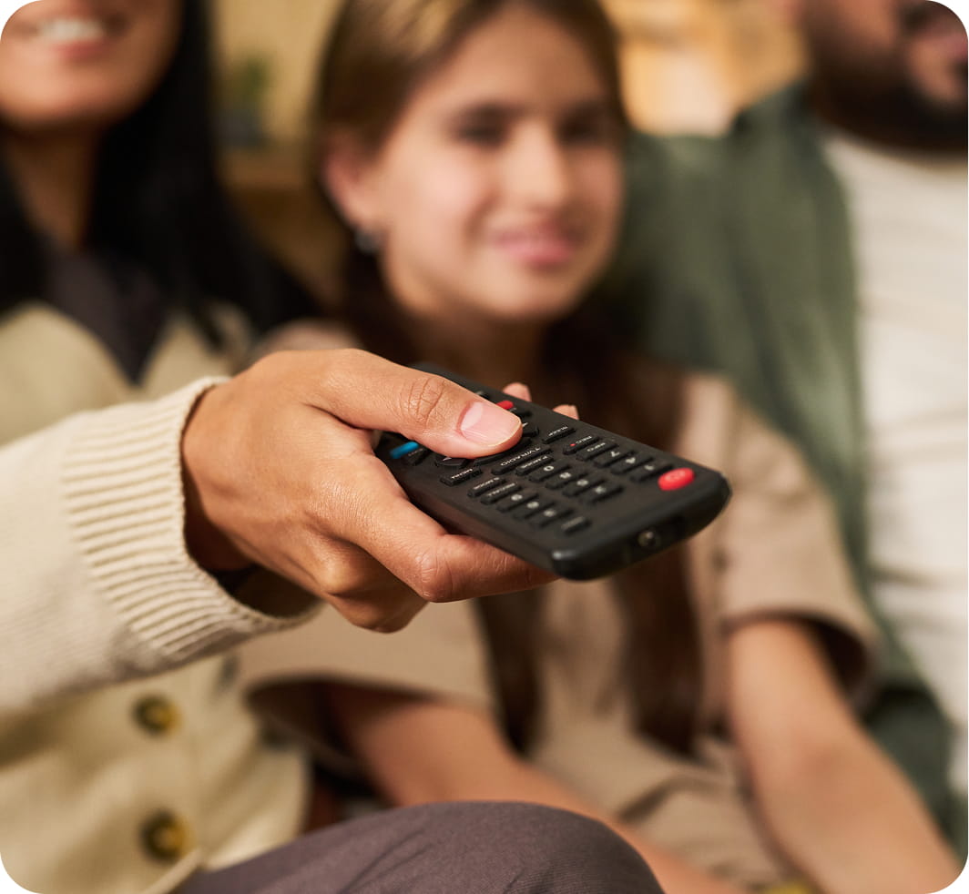 A close-up of a hand holding a TV remote, with a smiling family in soft focus in the background, conveying a cozy and relaxed atmosphere.