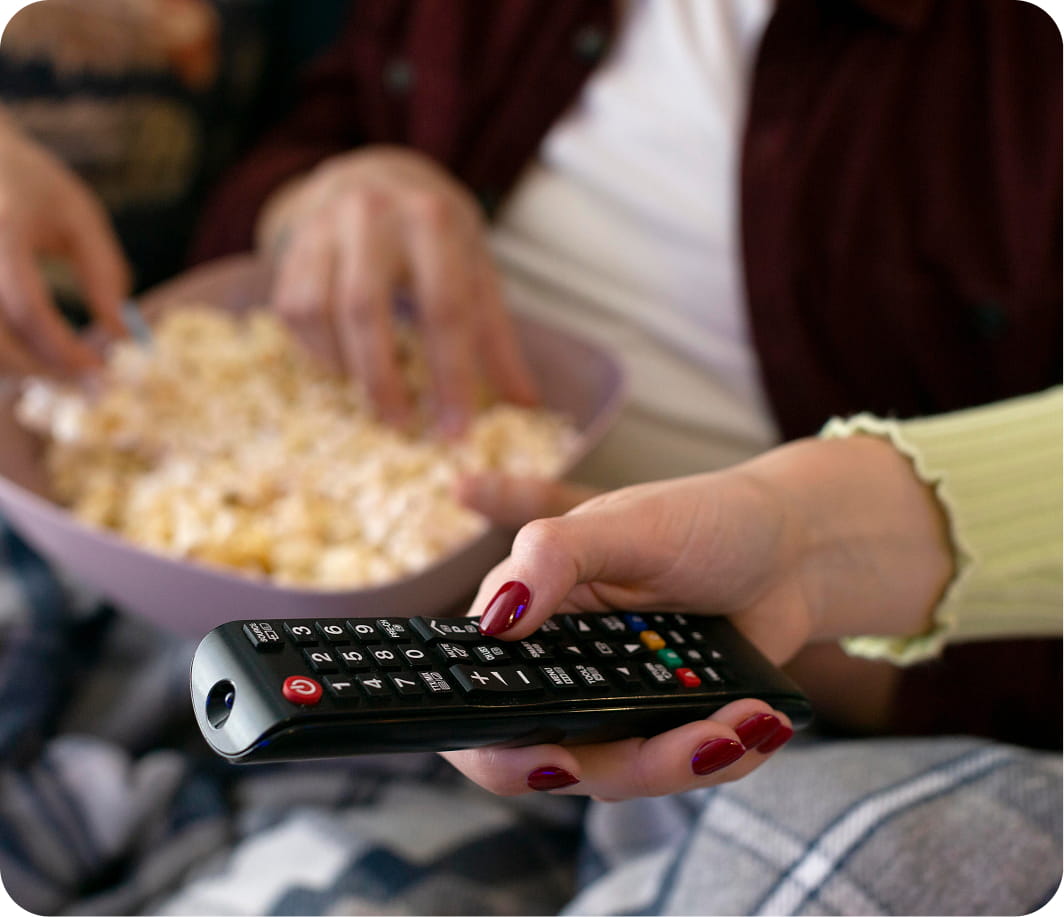 A hand with red nail polish holds a TV remote, while another hand reaches into a bowl of popcorn. The scene conveys relaxation and leisure.